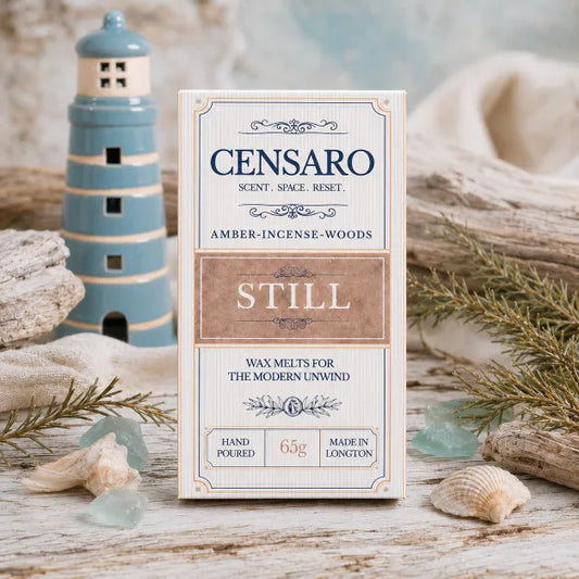 A boxed candle labeled CENSARO STILL stands upright on a rustic white surface with seashells, driftwood, and a small lighthouse decoration. The package is cream with navy text and a tan center panel, and the scene has a soft coastal styling.