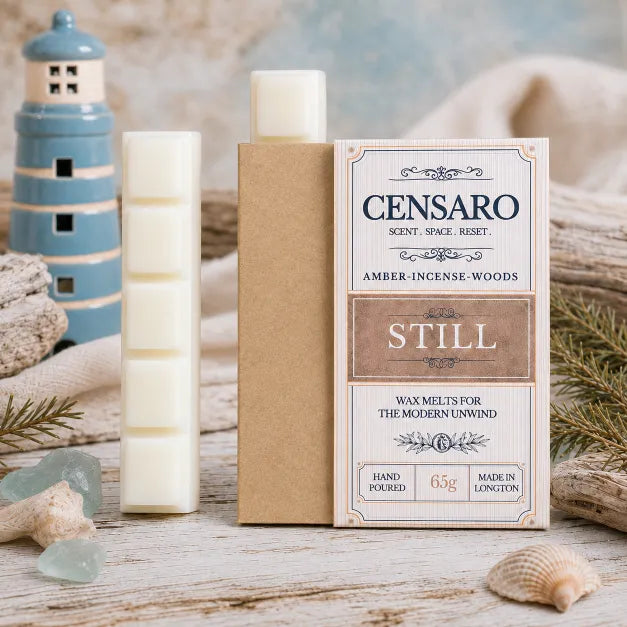 A wax melt product display featuring a kraft paper box labeled CENSARO STILL, a single white wax melt stick, and a decorative blue lighthouse prop on a rustic beach-themed surface.