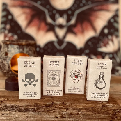 Four wax melts wrapped in vintage paper with spooky label designs and informative text.  With a blurred background of a bat and wax warmer on a rustic wooden surface.