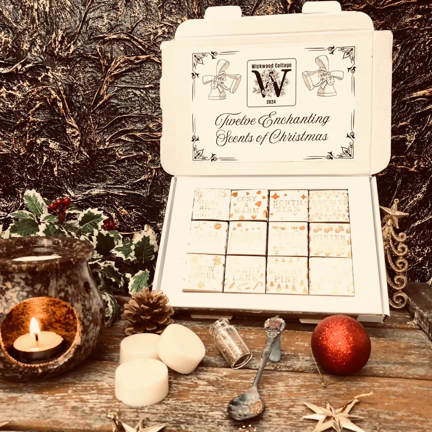 An open white Christmas box full of square individually wrapped wax melts in 12 different fragrances. Each melt is wrapped in white paper with gold text.  The box sits on a rustic wooden surface surrounded by holly, pinecones and a wax melt burner. Three individual wax melts discs sit Infront of the box along with a bauble, spoon and bottle full of glitter. 