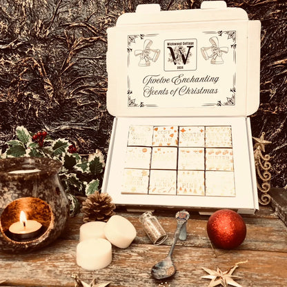 An open white Christmas box full of square individually wrapped wax melts in 12 different fragrances. Each melt is wrapped in white paper with gold text.  The box sits on a rustic wooden surface surrounded by holly, pinecones and a wax melt burner. Three individual wax melts discs sit Infront of the box along with a bauble, spoon and bottle full of glitter. 