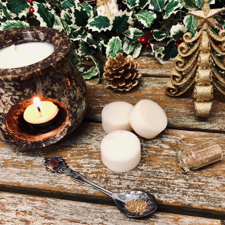An enchanted image showing three white wax melt discs, to the left is a rustic wax burner with a glowing candle full of melting wax. In the background is a bed of holly and to the right of the wax melts is a gold Christmas tree decoration. In the foreground is a decorative spoon full of glitter all sitting on a rustic wooden surface.