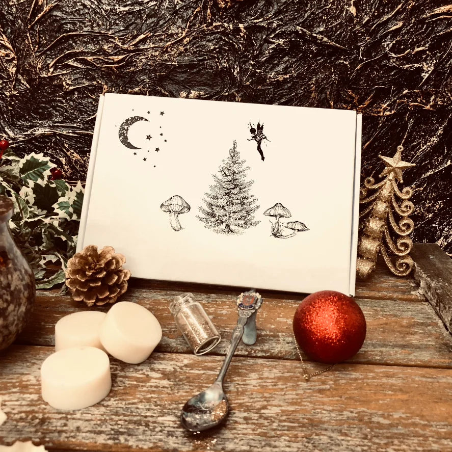 An enchanted image showing the front of our Christmas box of wax melts. Three white wax melt discs sit in front along with a pinecone, a decorative spoon full of glitter a small bottle filled with glitter and a red bauble. All sitting on a rustic wooden surface.