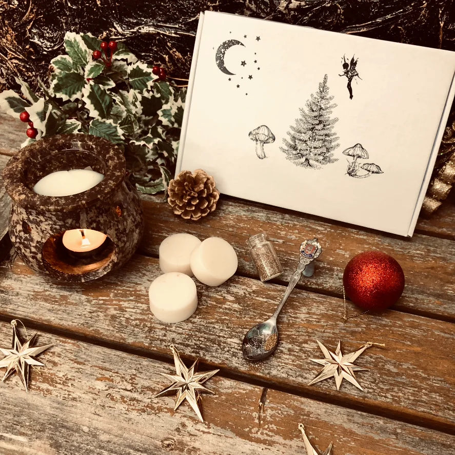 An enchanted image showing the front of our Christmas box of wax melts. Three white wax melt discs sit in front along with a pinecone, a decorative spoon full of glitter a small bottle filled with glitter and a red bauble.  To the left of the box is a mound of holly and a rustic looking wax burner with a glowing tealight and melting wax. All sitting on a rustic wooden surface.