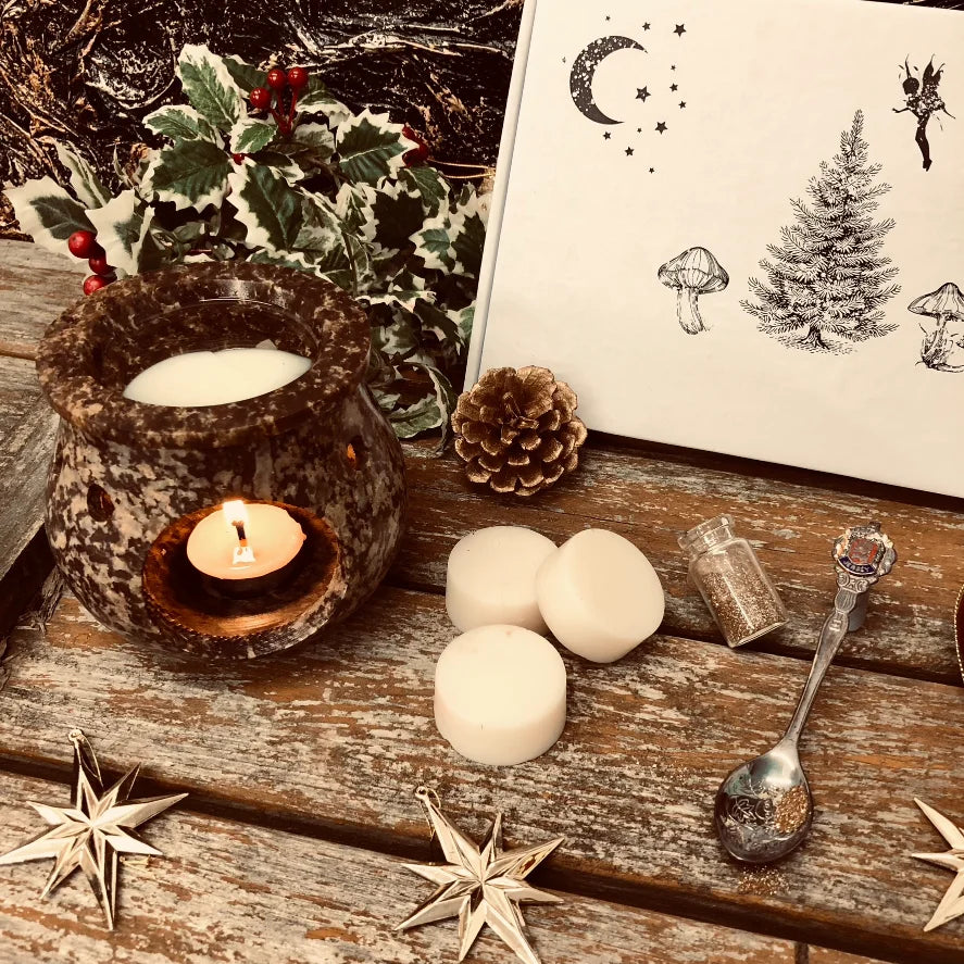 An enchanted image showing the front of our Christmas box of wax melts. A close-up view. Three white wax mel discs sit in front along with a pinecone, a decorative spoon full of glitter a small bottle filled with glitter and a red bauble. All sitting on a rustic wooden surface.
