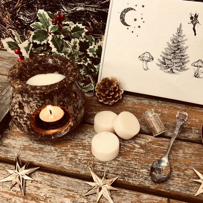 An enchanted image showing the front of our Christmas box of wax melts. A close-up view. Three white wax mel discs sit in front along with a pinecone, a decorative spoon full of glitter a small bottle filled with glitter and a red bauble. All sitting on a rustic wooden surface.