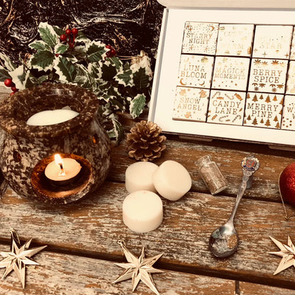 An enchanted image of an open white Christmas box full of square individually wrapped wax melts in 12 different fragrances. Each melt is wrapped in white paper with gold text.  The box sits on a rustic wooden surface to the left is a piece of holly and a pinecone.  A rustic looking wax warmer with a glowing tealight and melting wax.  In the foreground three white wax melt discs a spoon and a small bottle of glitter all resting on a rustic wooden surface.