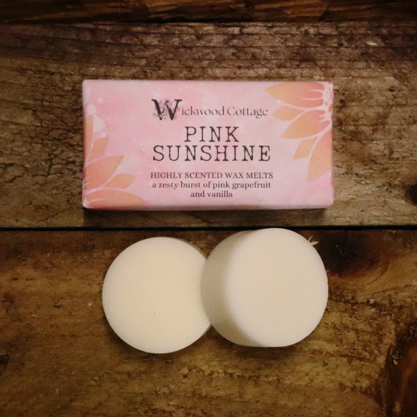 A rectangle paper package of 'Pink Sunshine' wax melts by Wickwood Cottage. The packaging is pink with sunflower patterns and descriptive text, displayed on top of a rustic wooden table. Below the pack are two wax melt discs displaying what's inside.