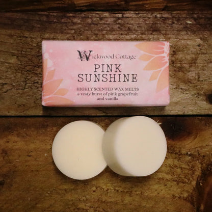 A rectangle paper package of 'Pink Sunshine' wax melts by Wickwood Cottage. The packaging is pink with sunflower patterns and descriptive text, displayed on top of a rustic wooden table. Below the pack are two wax melt discs displaying what's inside.