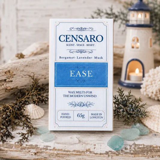 A wrapped fragrance product labeled CENSARO EASE, stands upright on a rustic white surface with decorative seashells, driftwood, greenery, and a lighthouse tea light in the background.