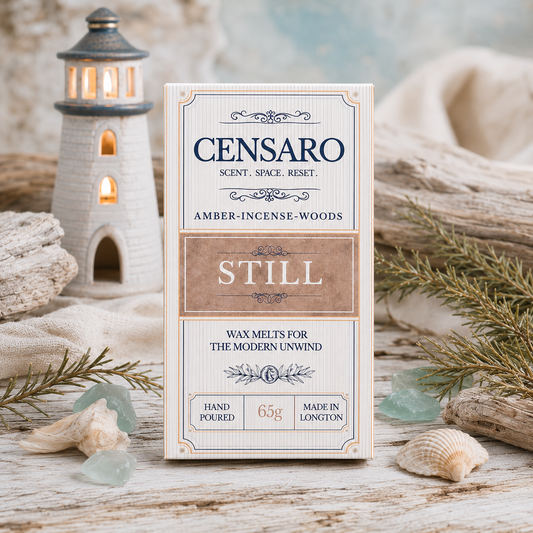 A wrapped fragrance product labeled CENSARO STILL, stands on a rustic white surface with seashells, driftwood, and a small lighthouse tea light. The wrapping is cream and tan with black text and reads Amber-Incense-Woods, Still, 65g, hand poured, and made in Longton.