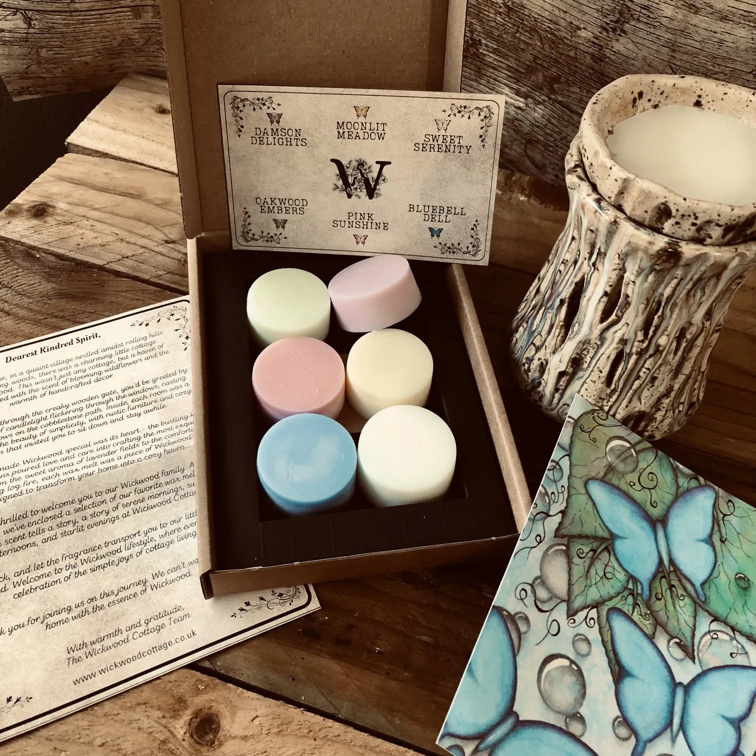 A box of colorful wax melt discs from Wickwood Cottage, with various shades including pink, blue, and white, displayed alongside a ceramic wax warmer, postcard and a printed story. Sitting on top of a rustic wooden surface.