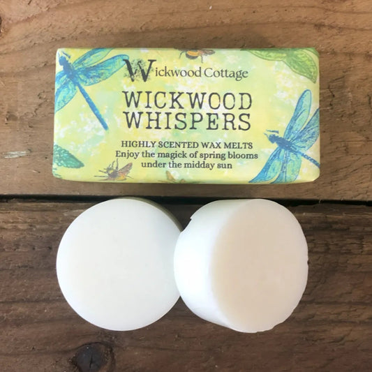 Two white wax melt discs displayed on a rustic wooden surface beneath a Wickwood Cottage label reading “Wickwood Whispers,” decorated with dragonflies and soft spring colours.