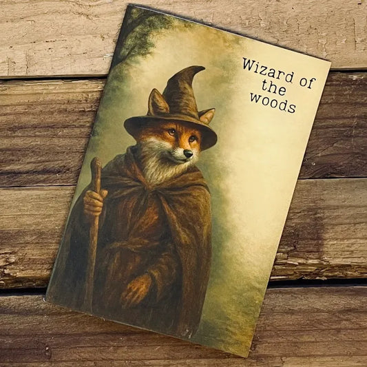 A rustic A5 notebook featuring a vintage-style illustration of a fox wizard in a brown cloak and pointed hat, holding a wooden staff in a misty forest. The cover reads “Wizard of the woods” and sits on a textured wooden background, giving the product a cosy, woodland, cottagecore aesthetic.