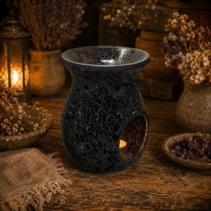 A dark, textured ceramic wax warmer with a crackle glaze and a cutout for a tea light, styled on a rustic wooden table with dried flowers and warm lanterns in the background.