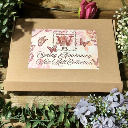 The front of the 'Spring Awakening' gift box set a postal box with a pink label displaying the Wickwood Cottage logo, name of the collection and decorated with butterflies and flowers. Surrounding the box an array of florals on a rustic wooden table.