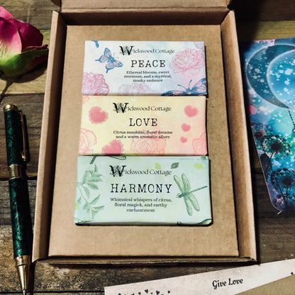 A collection of wax melts from Wickwood Cottage, three wax melts displayed in a box. Peace, Love and Harmony. The wax melts are in various colors and have labels with different scent names. To the side of the box is a pen, rose and art postcard. Sitting on top of a rustic table.