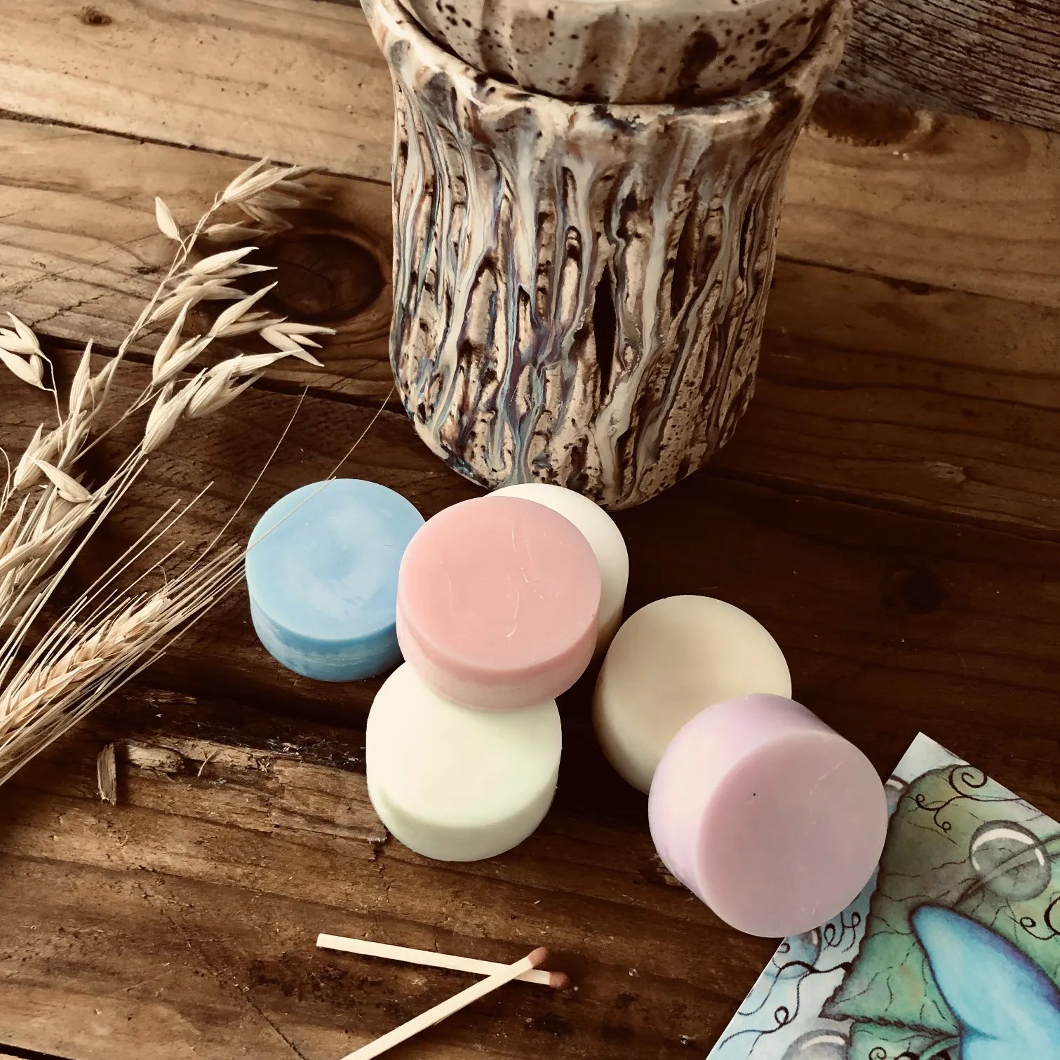 Six colorful wax melt discs from Wickwood Cottage, with various shades including pink, blue, and white, displayed alongside a ceramic wax warmer, postcard and a printed story. Sitting on top of a rustic wooden surface.