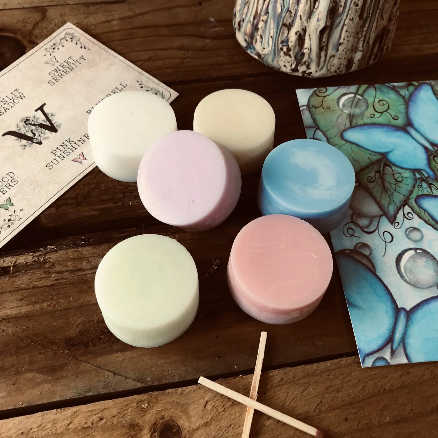 Six colorful wax melt discs from Wickwood Cottage, with various shades including pink, blue, and white, displayed alongside a ceramic wax warmer, matches, postcard and a printed name card. Sitting on top of a rustic wooden surface.