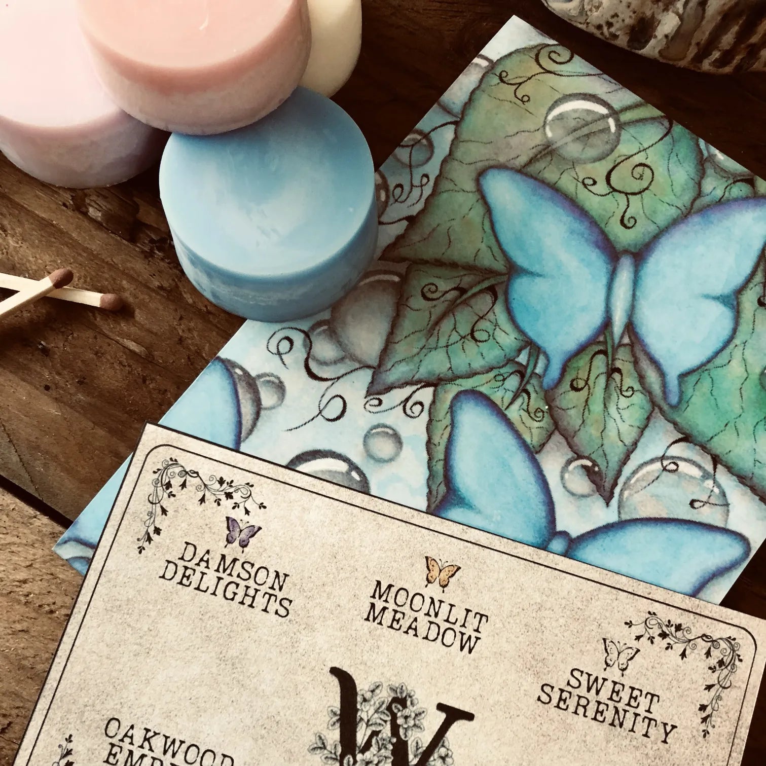 Colorful wax melt discs from Wickwood Cottage, with various shades including pink, blue, and white, displayed alongside a ceramic wax warmer, matches, postcard and a printed name card. Sitting on top of a rustic wooden surface.