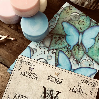 Colorful wax melt discs from Wickwood Cottage, with various shades including pink, blue, and white, displayed alongside a ceramic wax warmer, matches, postcard and a printed name card. Sitting on top of a rustic wooden surface.