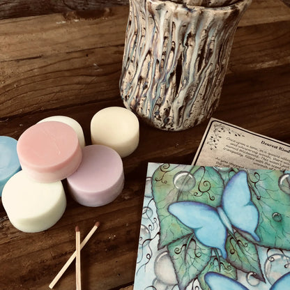 Six colorful wax melt discs from Wickwood Cottage, with various shades including pink, blue, and white, displayed alongside a ceramic wax warmer, matches, postcard and a printed name card. Sitting on top of a rustic wooden surface.