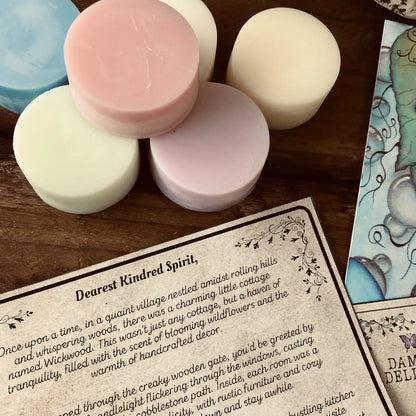 Six colorful wax melt discs from Wickwood Cottage, with various shades including pink, blue, and white, displayed alongside a postcard and a printed story. Sitting on top of a rustic wooden surface.