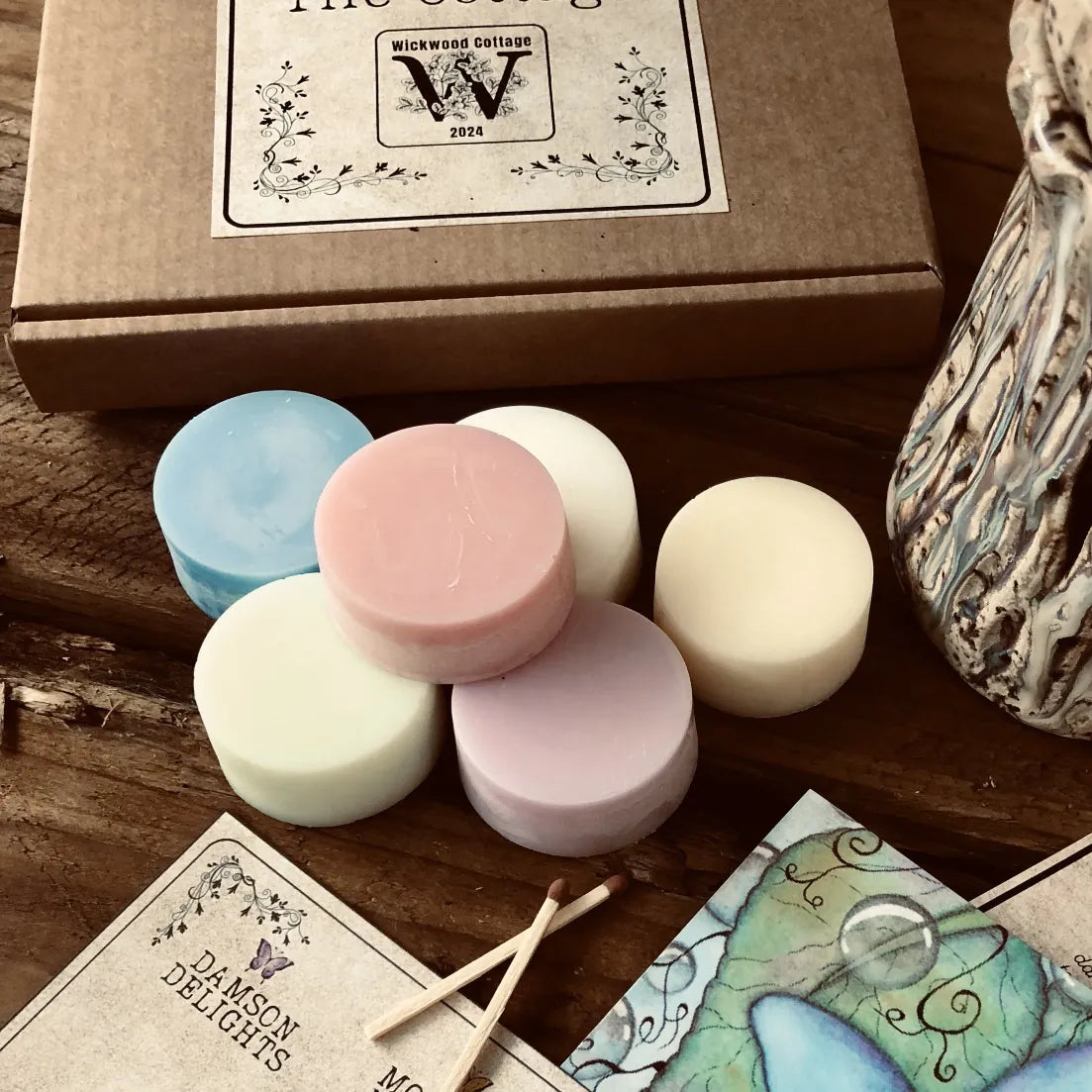 A box showing the front label, in front are colorful wax melt discs from Wickwood Cottage, with various shades including pink, blue, and white, displayed alongside a ceramic wax warmer, postcard and a printed story. Sitting on top of a rustic wooden surface.