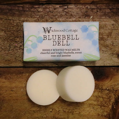A rectangle paper package of 'Bluebell Dell' wax melts by Wickwood Cottage. The packaging is pale blue with bluebell patterns and descriptive text, displayed on a rustic wooden table. Underneath there are two wax melt discs showing what's inside.