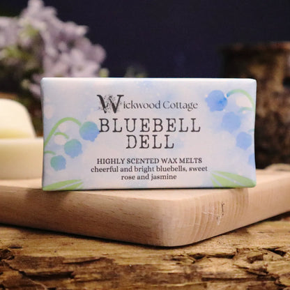 A rectangle paper package of 'Bluebell Dell' wax melts by Wickwood Cottage. The packaging is pale blue with bluebell patterns and descriptive text, displayed on top of a wood platter on a rustic wooden table. Behind is a blurred background of flowers and a wax warmer.