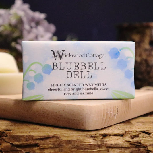 A rectangle paper package of 'Bluebell Dell' wax melts by Wickwood Cottage. The packaging is pale blue with bluebell patterns and descriptive text, displayed on top of a wood platter on a rustic wooden table. Behind is a blurred background of flowers and a wax warmer.