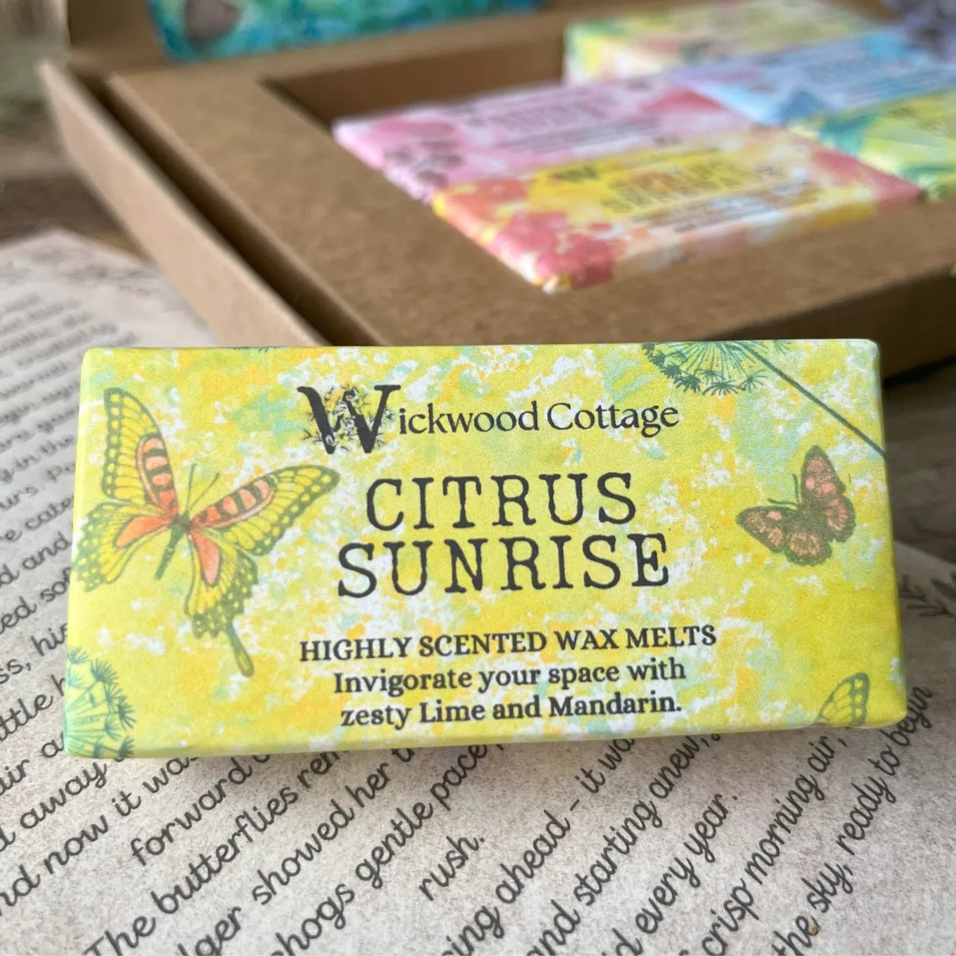 Paper packaging of Wickwood Cottage Citrus Sunrise highly scented wax melts. The packaging has a butterfly pattern with yellow background and informative text. In the background is the gift box. Underneath is the story which comes with the gift box.