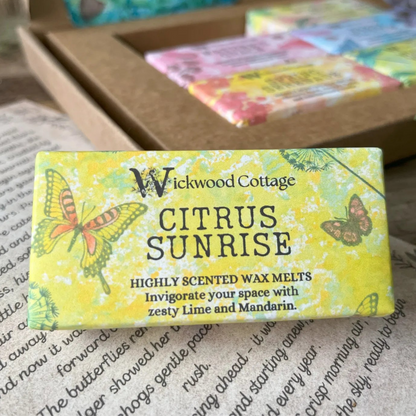 Paper packaging of Wickwood Cottage Citrus Sunrise highly scented wax melts. The packaging has a butterfly pattern with yellow background and informative text. In the background is the gift box. Underneath is the story which comes with the gift box.