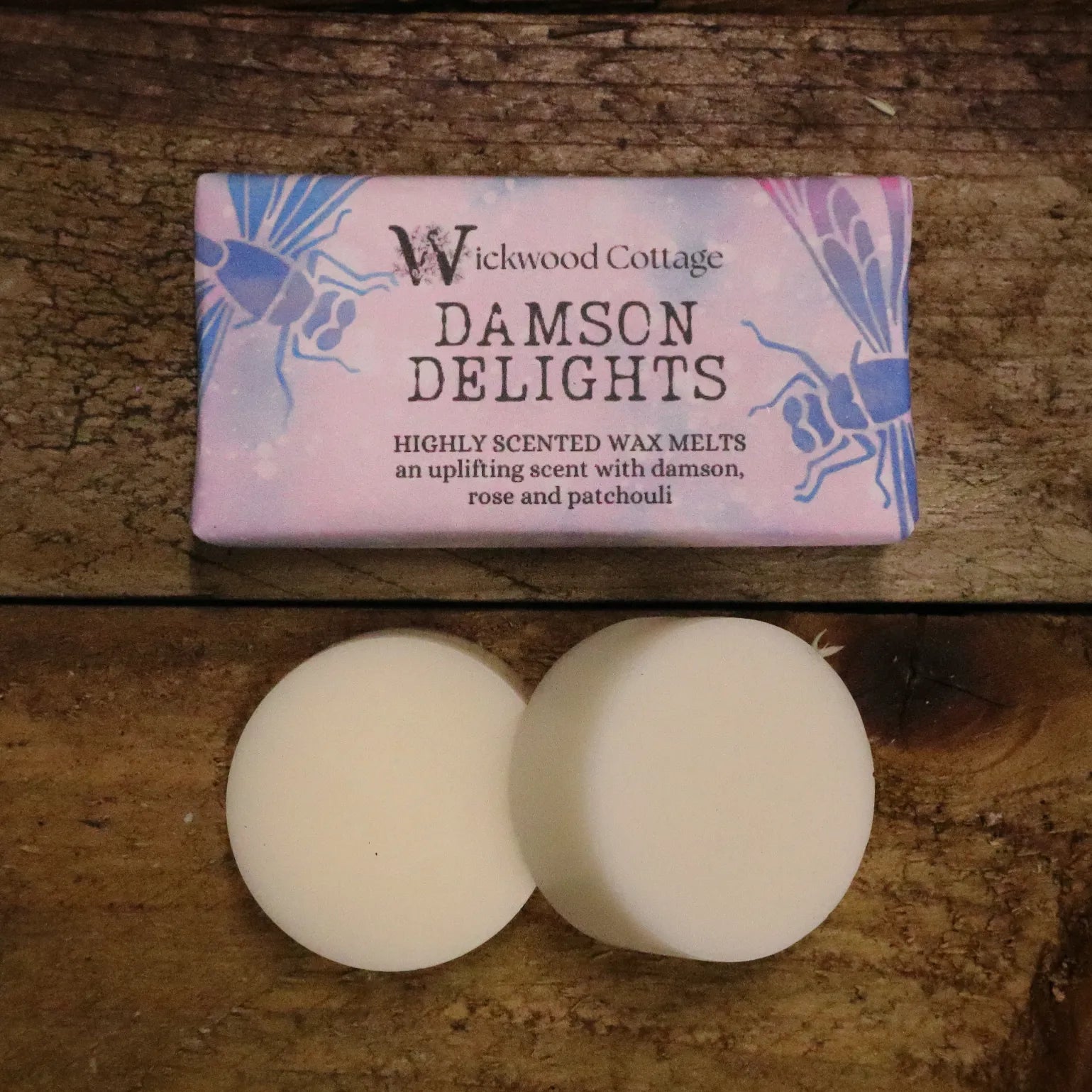 A rectangle paper package of 'Damson Delights' wax melts by Wickwood Cottage, featuring highly scented notes of damson, rose, and patchouli. The packaging is pink with dragon fly patterns. Below the package is an example of what's inside, two 20g wax melt discs and the product is displayed on a rustic wooden table. 