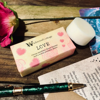 Paper packaging of Wickwood Cottage Love highly scented wax melts. The packaging has pink hearts and a floral pattern with yellow background. The image is set within a rustic table depicting a loved one's letter on the table a single rose a pen and the story and postcard.  