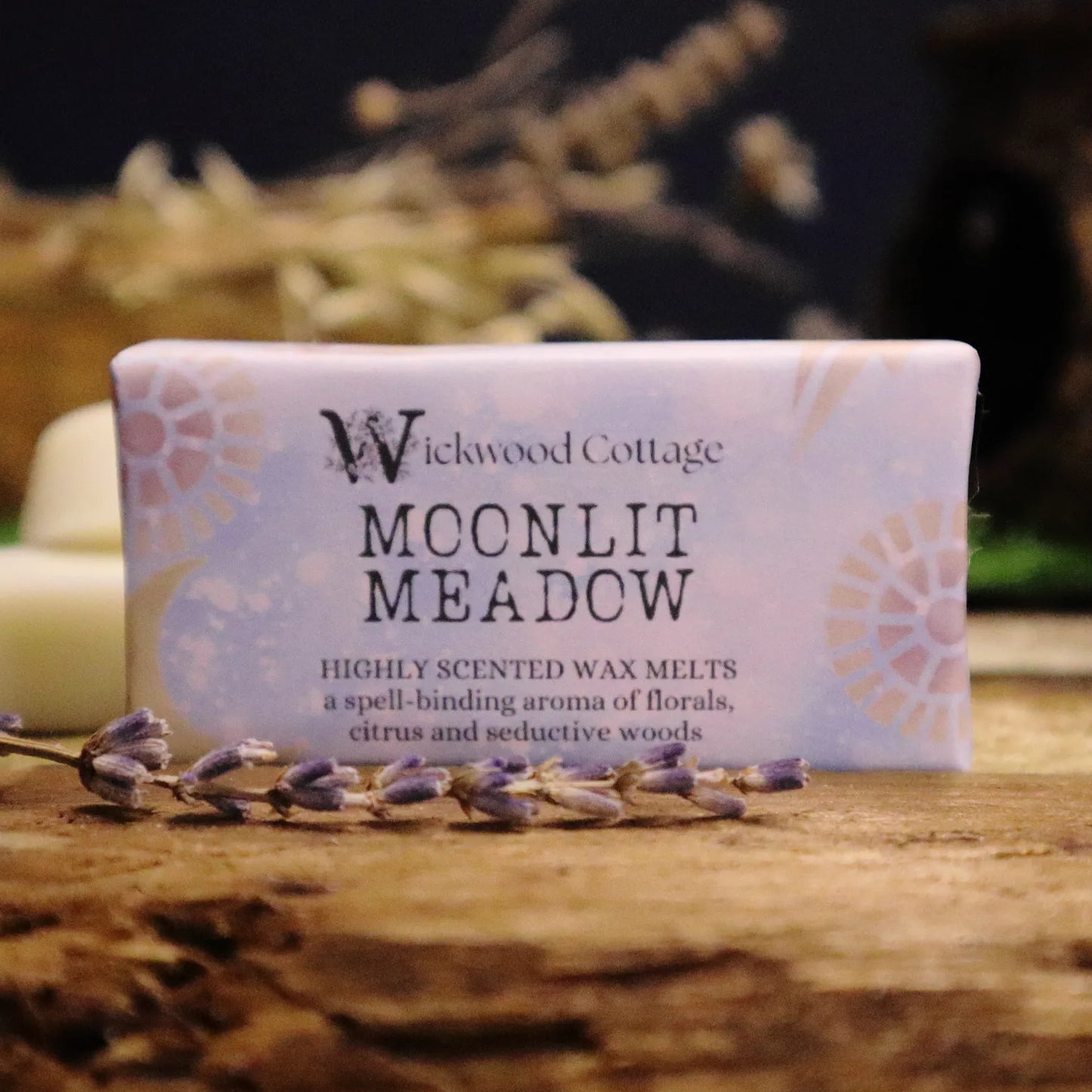 A rectangle paper package of 'Moonlit Meadow' wax melts by Wickwood Cottage. The packaging is lilac with a floral pattern and descriptive text, displayed on top of a rustic wooden table. Behind is a blurred background of dried flowers and a wax warmer. 