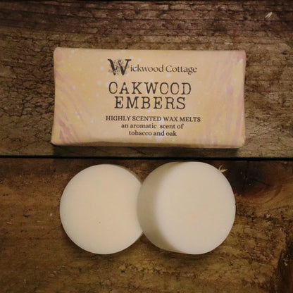 A rectangle paper package of 'Oakwood Embers' wax melts by Wickwood Cottage. The packaging is light brown with a tree bark pattern and descriptive text, displayed on top of a rustic wooden table. Below the package are two wax melt discs showing the contents.