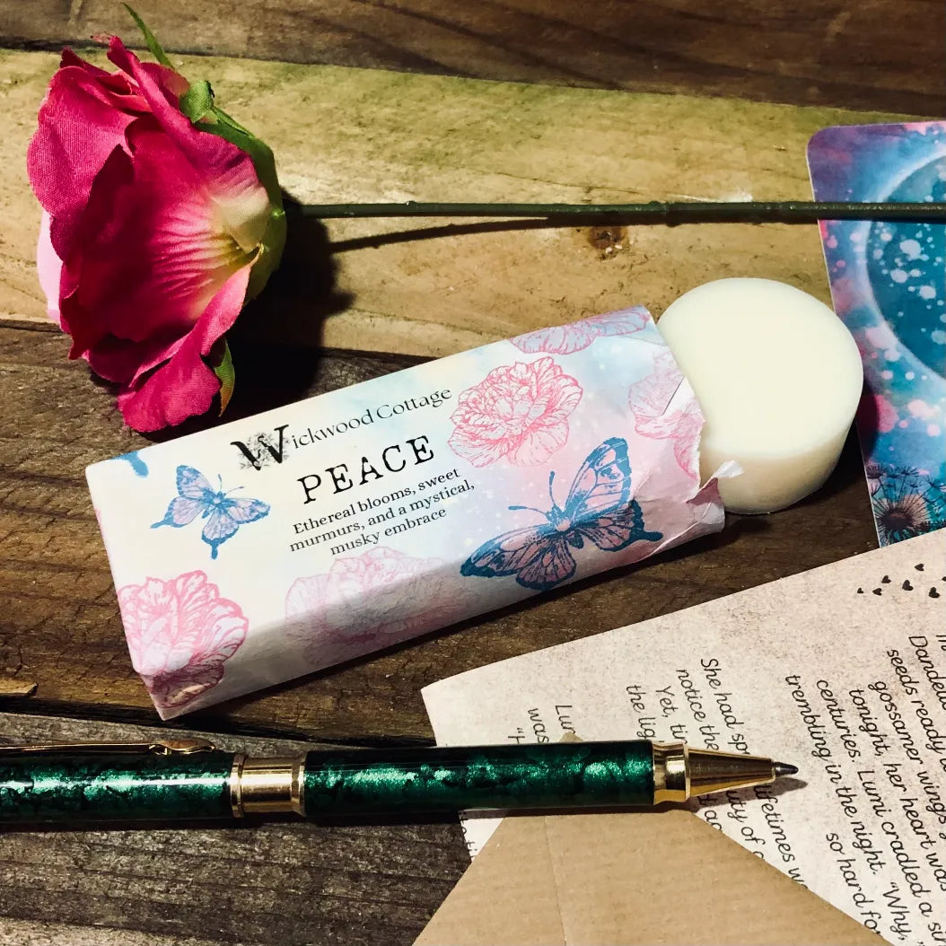 Paper packaging of Wickwood Cottage Peace highly scented wax melts. The packaging has butterfly's and a floral pattern with lilac background. The image is set within a rustic table depicting a loved one's letter on the table a single rose a pen and the story and postcard.  