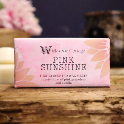 A rectangle paper package of 'Pink Sunshine' wax melts by Wickwood Cottage. The packaging is pink with sunflower patterns and descriptive text, displayed on top of a rustic wooden table. The background is blurred with a brown wax warmer and a basket of pink flowers.