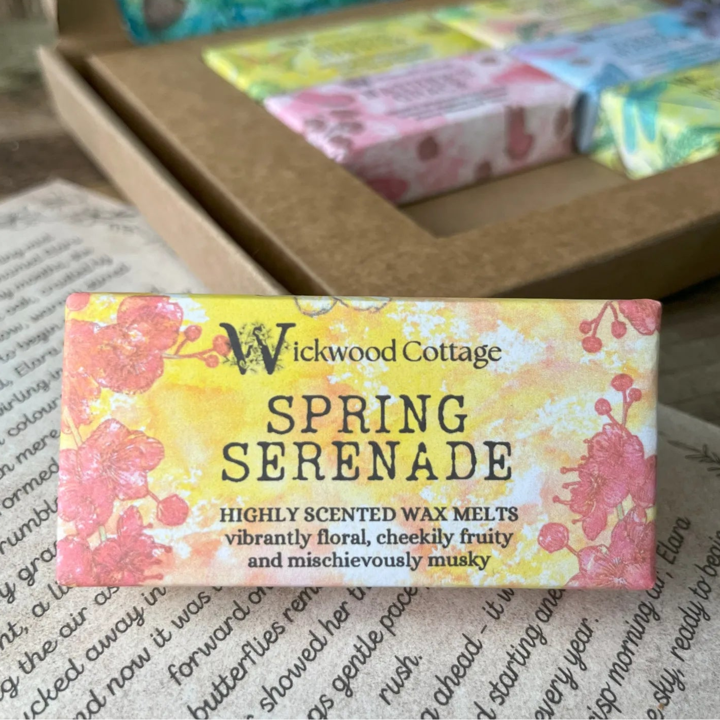 Paper packaging of Wickwood Cottage Spring Serenade highly scented wax melts. The packaging has a red floral pattern with yellow background and informative text. In the background is the gift box. Underneath is the story which comes with the gift box.