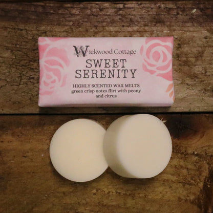 A rectangle paper package of 'Sweet Serenity' wax melts by Wickwood Cottage. The packaging is pink with a floral pattern and descriptive text, displayed on top of a rustic wooden table. Below is an example of its contents, two wax melt discs