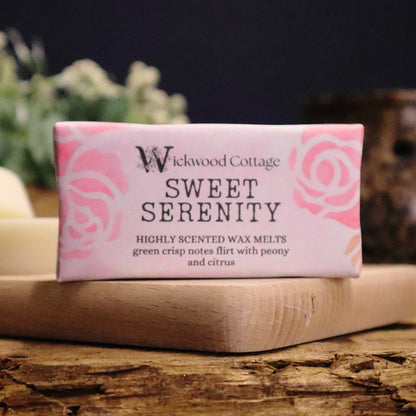 A rectangle paper package of 'Sweet Serenity' wax melts by Wickwood Cottage. The packaging is pink with a floral pattern and descriptive text, displayed on top of wood platter on a rustic wooden table. Behind is a blurred background of flowers and a wax warmer. 