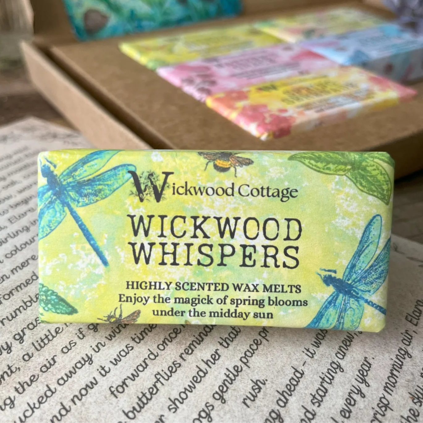 Paper packaging of Wickwood Cottage Wickwood Whispers highly scented wax melts. The packaging has a dragonfly pattern with yellow background and informative text. In the background is the gift box. Underneath is the story which comes with the gift box.