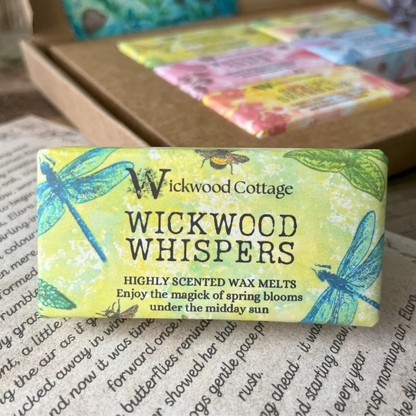 Paper packaging of Wickwood Cottage Wickwood Whispers highly scented wax melts. The packaging has a dragonfly pattern with yellow background and informative text. In the background is the gift box. Underneath is the story which comes with the gift box.