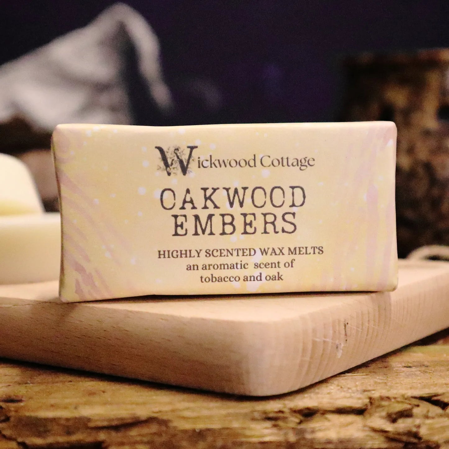 A rectangle paper package of 'Oakwood Embers' wax melts by Wickwood Cottage. The packaging is light brown with a tree bark pattern and descriptive text, displayed on top of a wood platter on a rustic wooden table. Behind is a blurred background of a wizard's hat and a wax warmer.